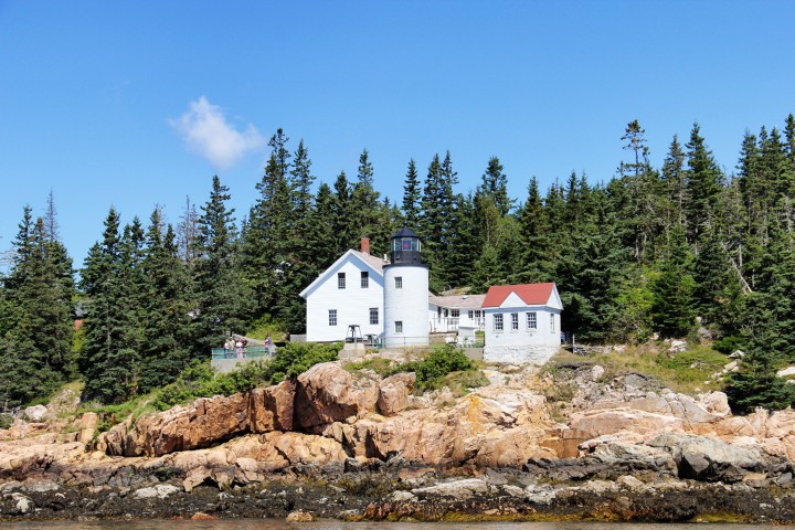 As seen from the ocean, the lighthouse encompassed by the red granite cliffs, imposing pines, and blue sky makes for a striking scene.